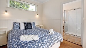A bedroom with a bed, a bedside table, a nightstand, a window with blinds, and a shower area. - ST PAULS PINK BAYSIDE SELF CONTAINED COTTAGE IN SORRENTO (Sorrento)