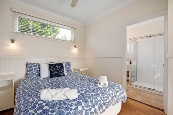 Un dormitorio con cama, mesita de noche, persianas en la ventana y zona de ducha.