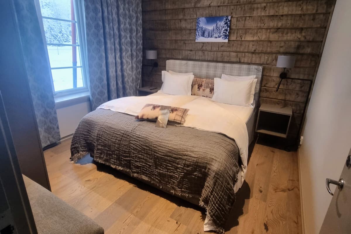 A bedroom with a wooden headboard, a bed with white linens, two bedside tables with lamps, and a window with curtains.