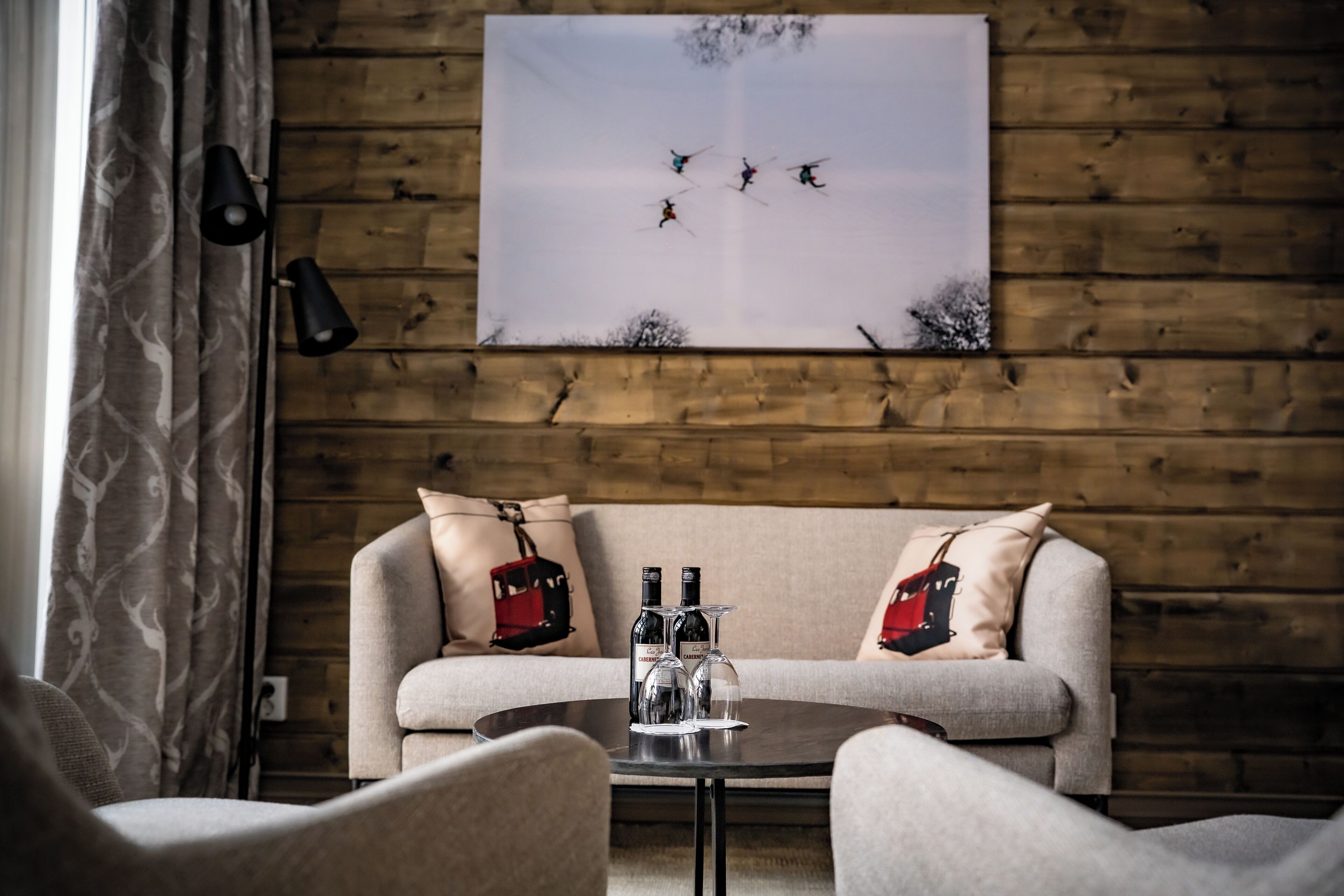 Um quarto acolhedor com parede de madeira, um sofá com almofadas estampadas, uma mesa redonda com garrafas de vinho e um quadro de esquiadores.