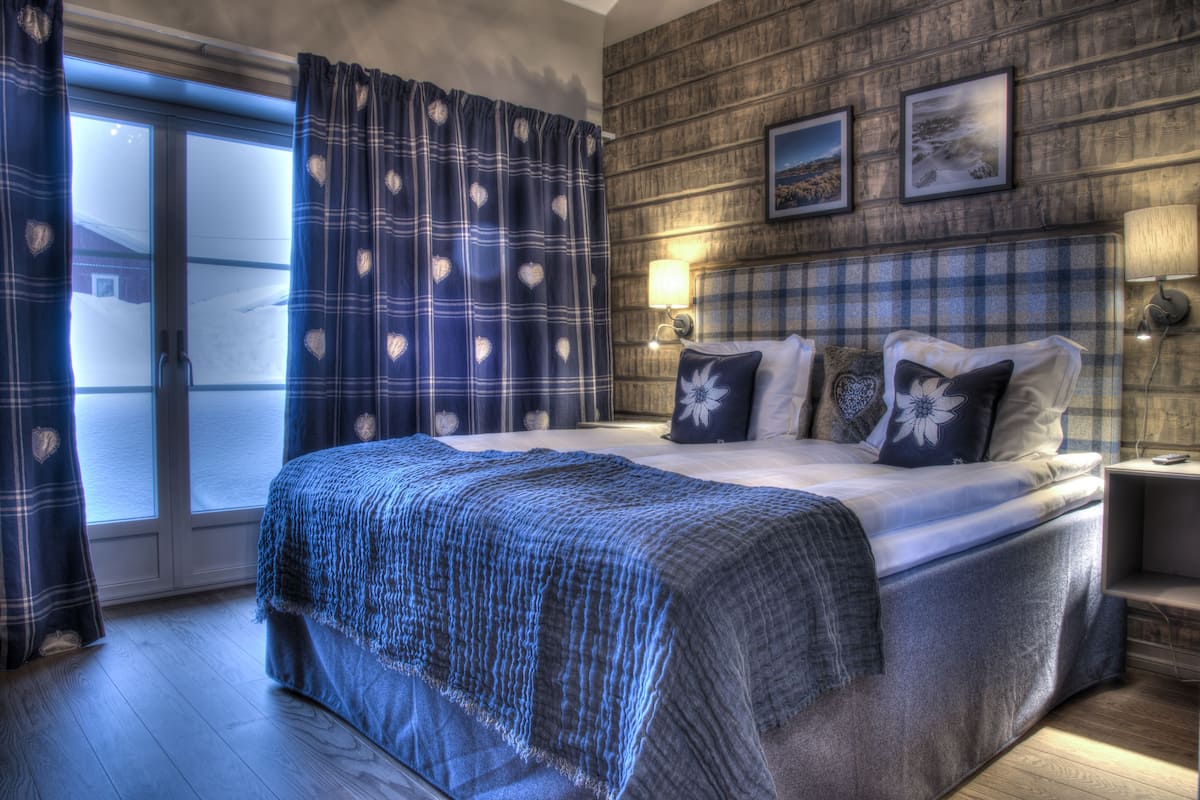 A bedroom with a large bed, a wooden headboard, a window with curtains, and a wall-mounted lamp.