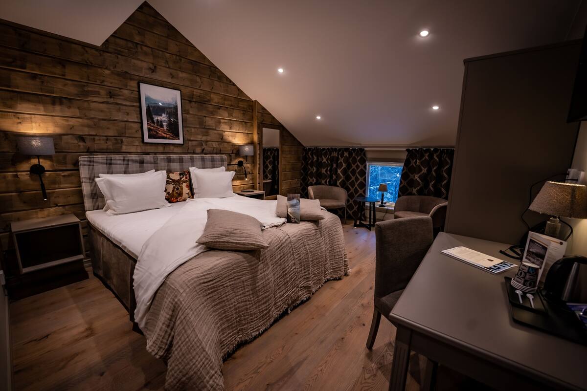 A bedroom with a wooden sloped ceiling, a bed with white linens, a desk with a lamp, and a window with curtains.