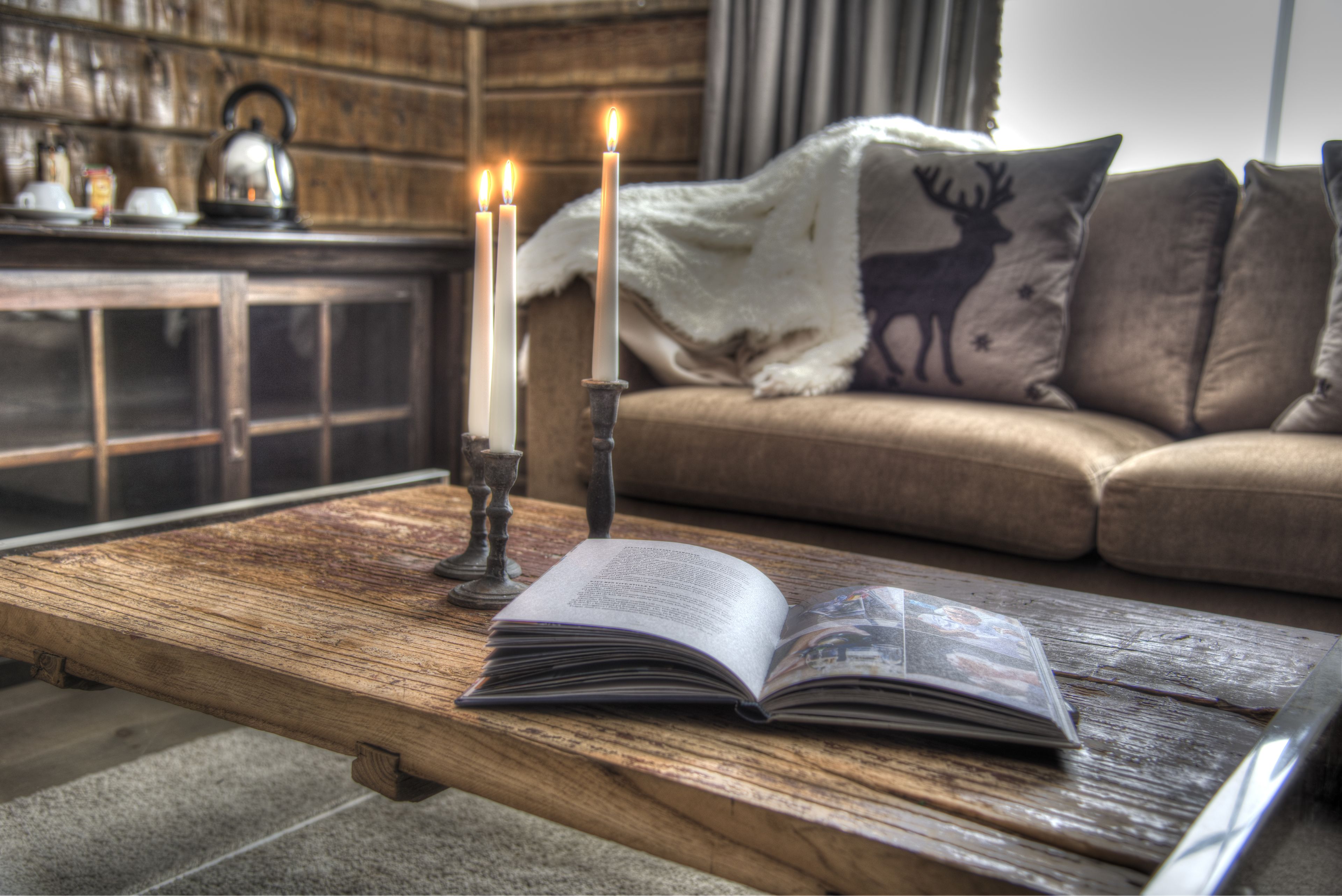A wooden coffee table with an open book and two lit candles. A sofa with a deer pillow is in the background.