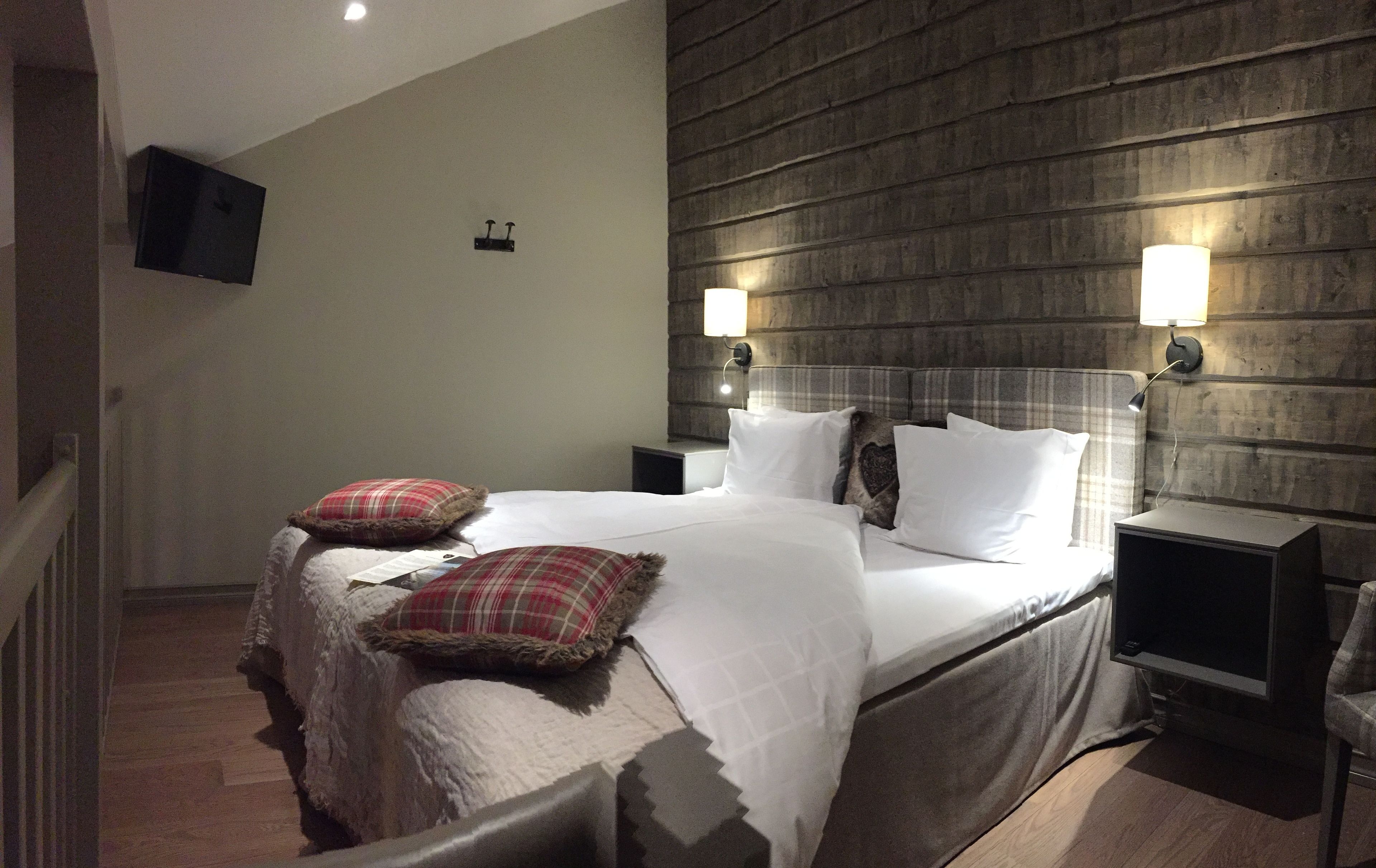 Una cama bien hecha con sábanas blancas y almohadas de cuadros, una mesita de noche con lámpara, y un televisor de pantalla plana montado en la pared.
