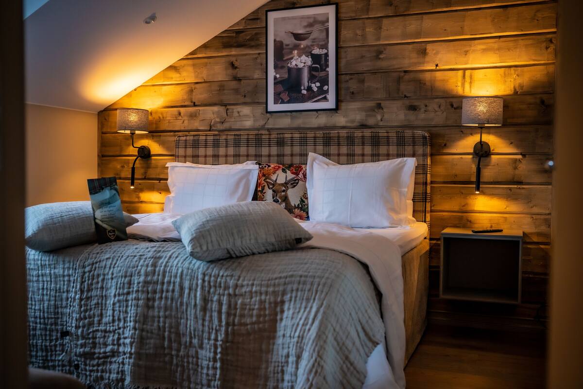 A bedroom with a wooden headboard, two bedside lamps, a framed picture above the bed, and a plaid-patterned pillow.
