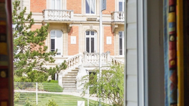 A view through a window of a large, historic building with multiple stories, a prominent entrance, and a staircase leading to the main door.