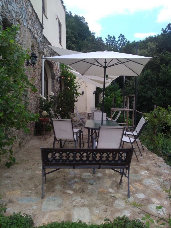 Une terrasse extérieure avec un sol en pierre, une table entourée de chaises et un grand parasol pour faire de l’ombre.