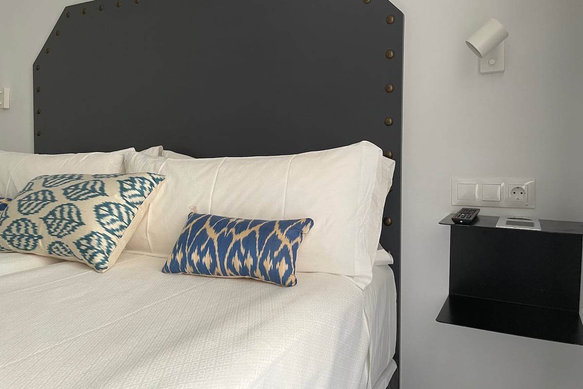 A bed with white bedding and patterned pillows, a dark headboard with rivets, a bedside table with a remote control, and a wall socket.
