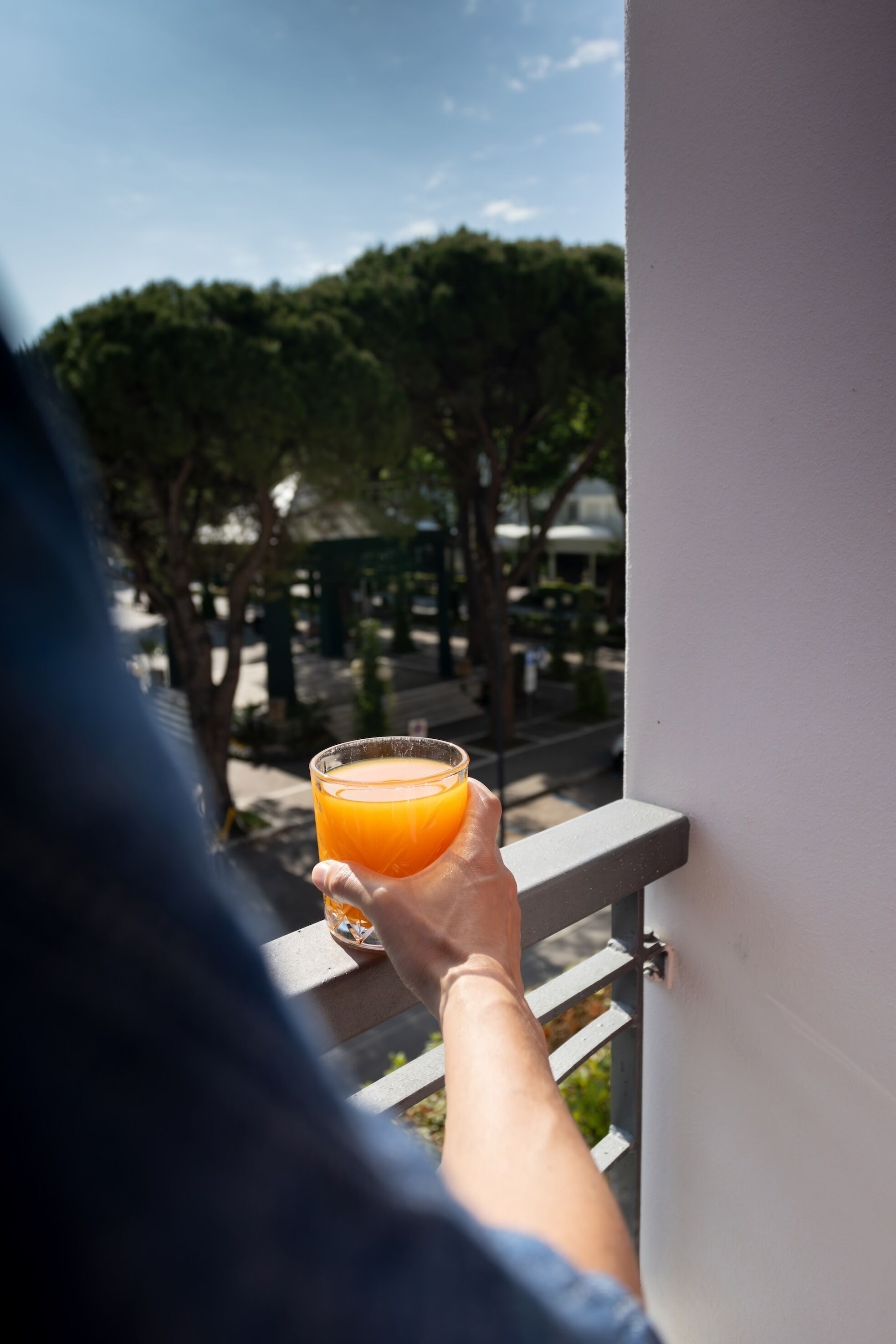 A person holding a glass of orange juice on a balcony with a railing, trees, and a building in the background.