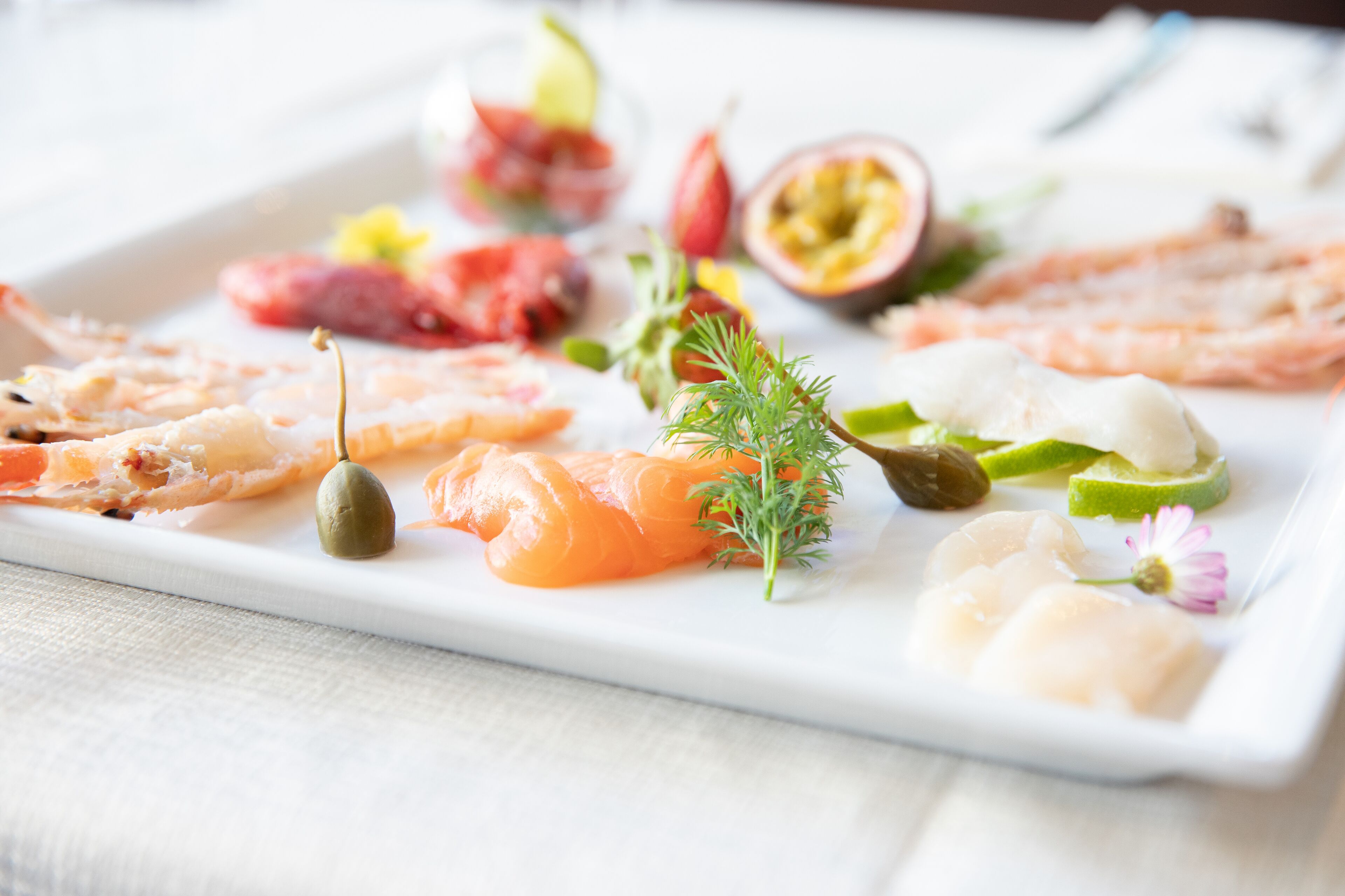 Variedad de mariscos dispuestos en un plato blanco rectangular, que incluyen lonchas de salmón, camarones y otros pescados, adornados con hierbas y pequeñas verduras.