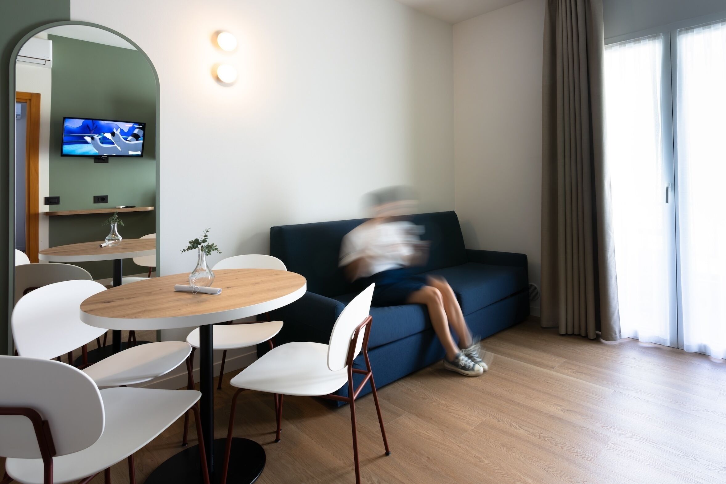 Una sala de estar moderna con una mesa redonda de madera, sillas blancas, un sofá azul oscuro y un televisor de pantalla plana montado en la pared.