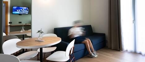Una sala de estar moderna con una mesa redonda de madera, sillas blancas, un sofá azul oscuro y un televisor de pantalla plana montado en la pared.