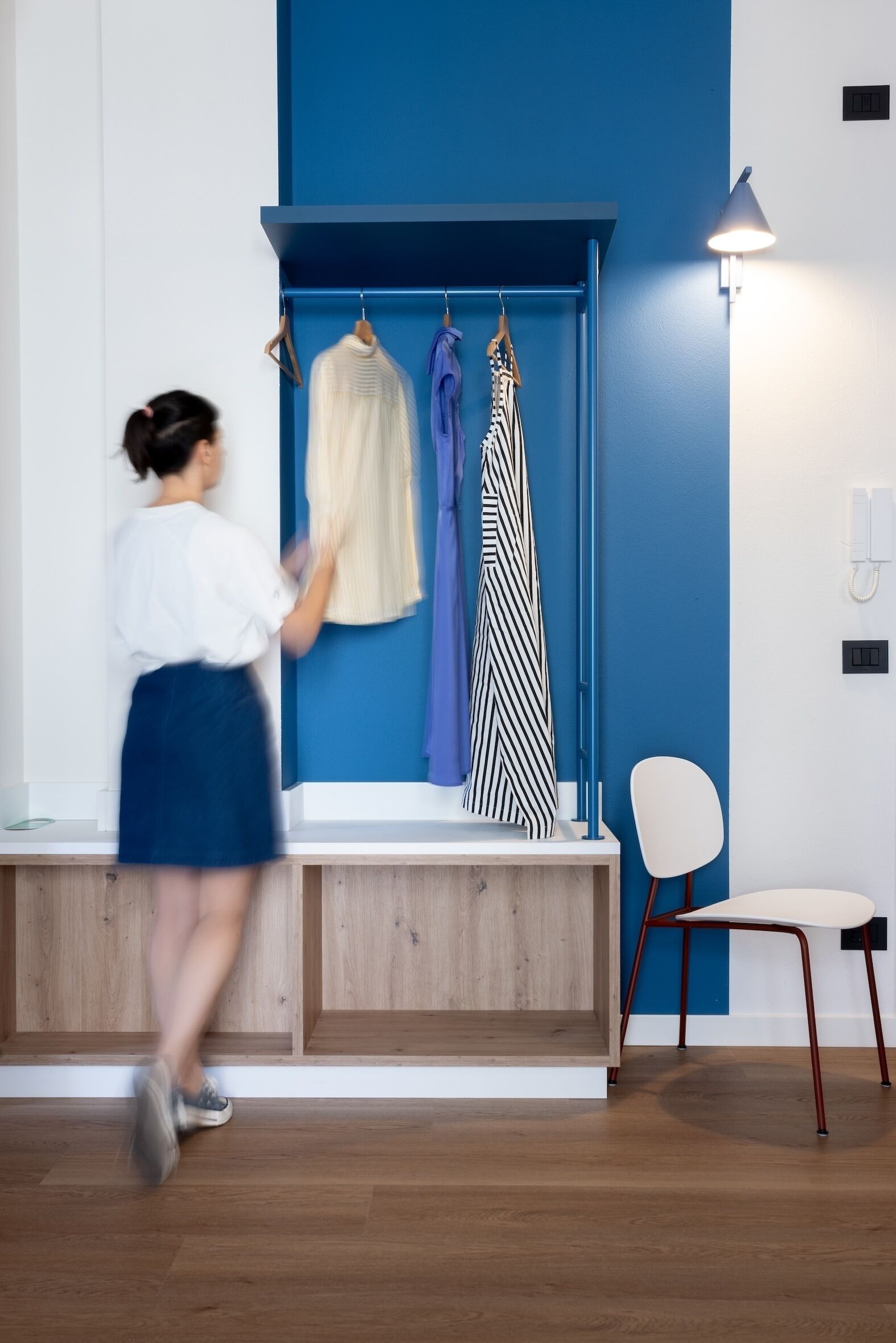 Une pièce avec un mur d’accent bleu, un banc en bois, une chaise blanche et un portant à vêtements sur lequel sont disposés différents articles d’habillement.