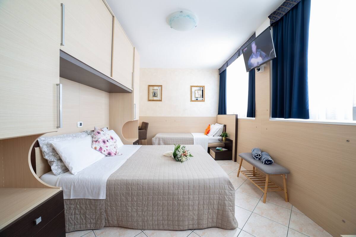 Una camera d'albergo con un letto grande, un letto più piccolo, una panca, una televisione e una finestra con tende.