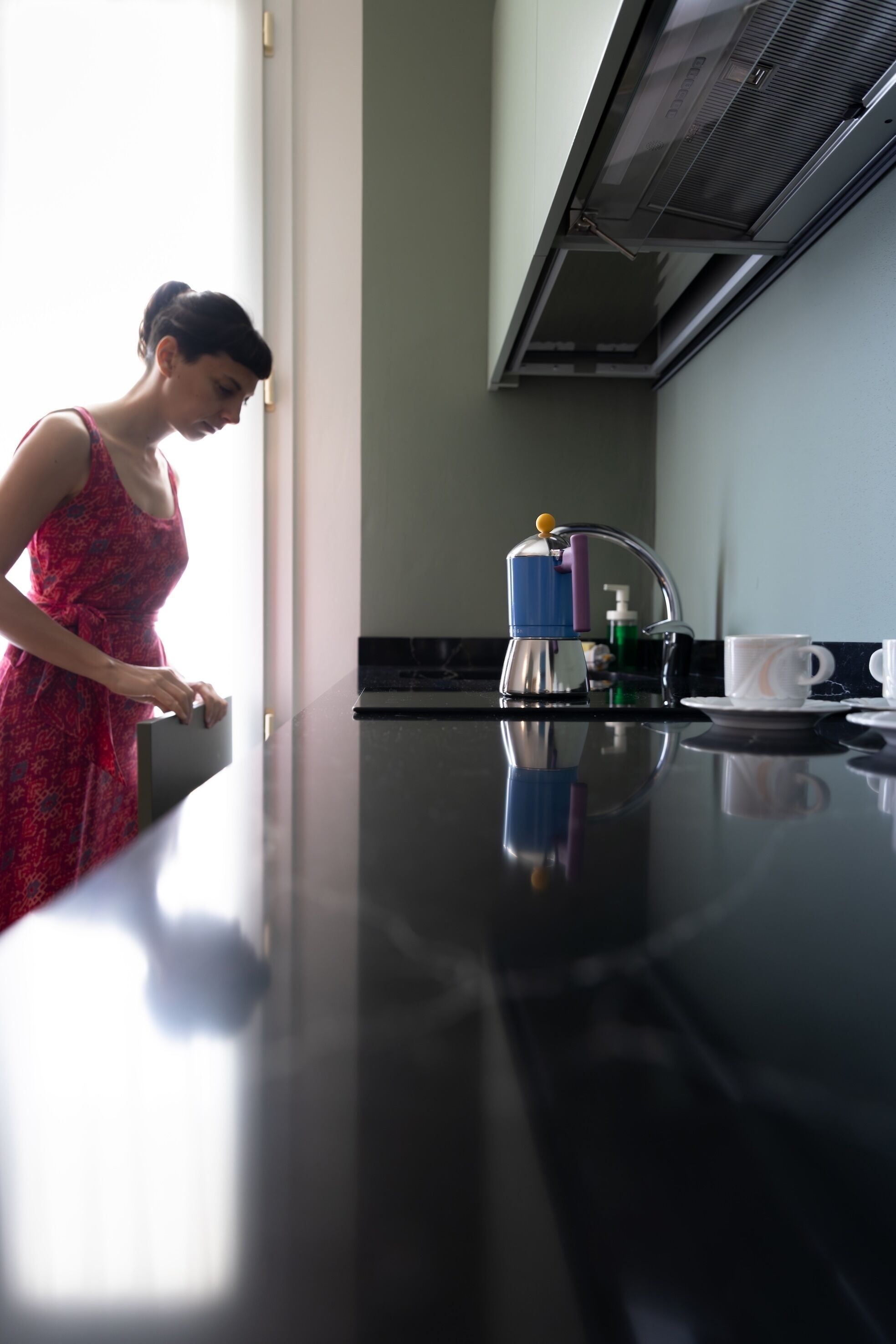 A woman in a red dress standing by a kitchen counter with a coffee maker, a cup, and a bottle of hand soap.