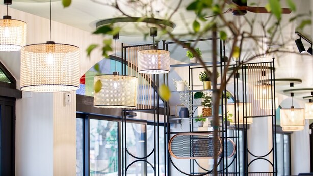 Ein modernes Interieur mit mehreren hängenden Pendelleuchten, einem Metallregal mit Pflanzen und einem großen Fenster, das viel Tageslicht hereinlässt.