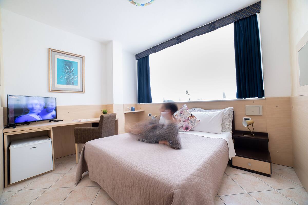 Una camera d'albergo con un letto, una scrivania con televisore, un piccolo frigorifero e una finestra con tende.