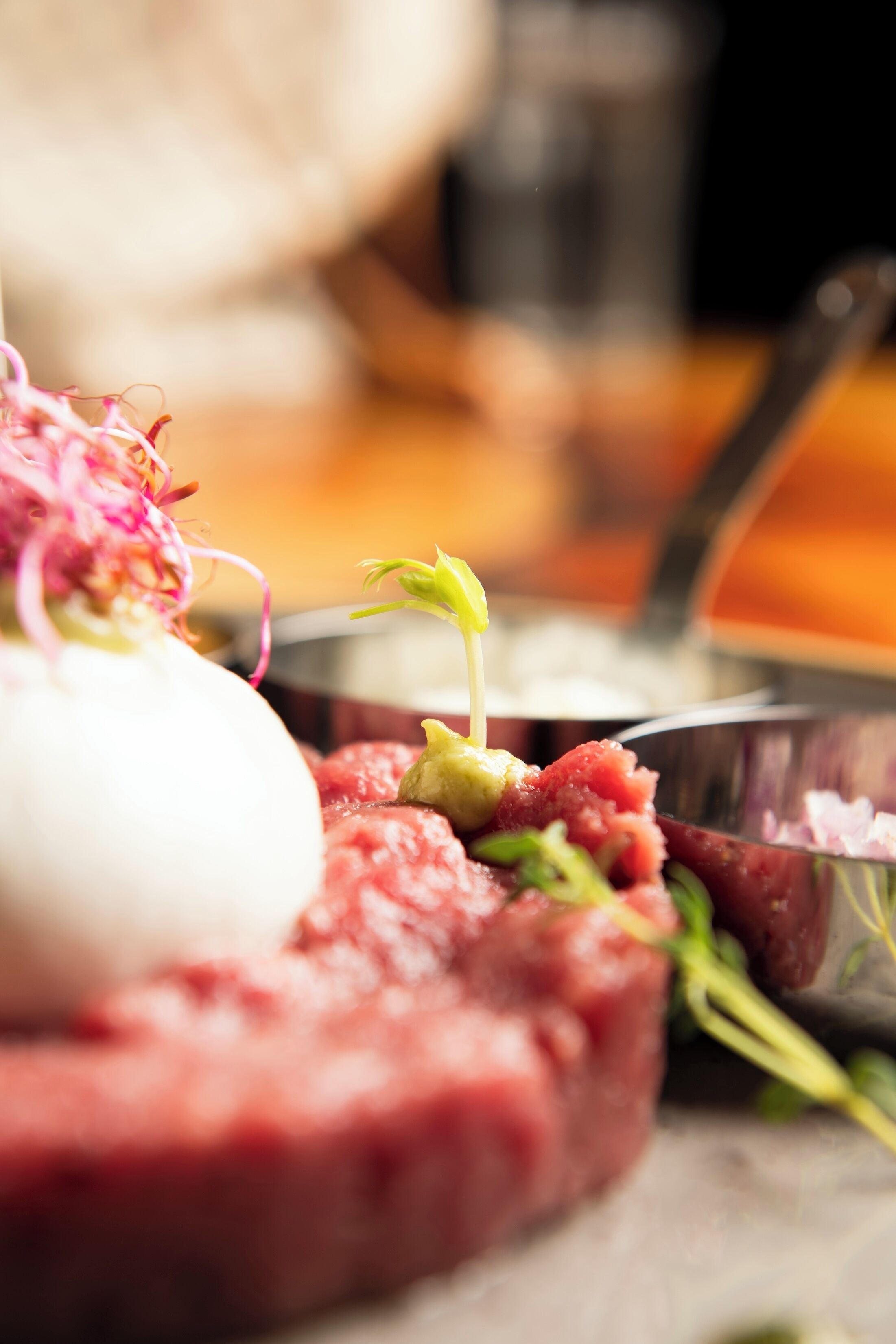 A close-up of a dish with a raw meat centerpiece, garnished with green herbs and a white spherical item, possibly cheese, on a metal plate.