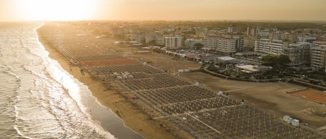Une vue aérienne d’une plage bordée de rangées de parasols et de chaises longues, à côté d’un paysage urbain avec des bâtiments et un ciel dégagé.