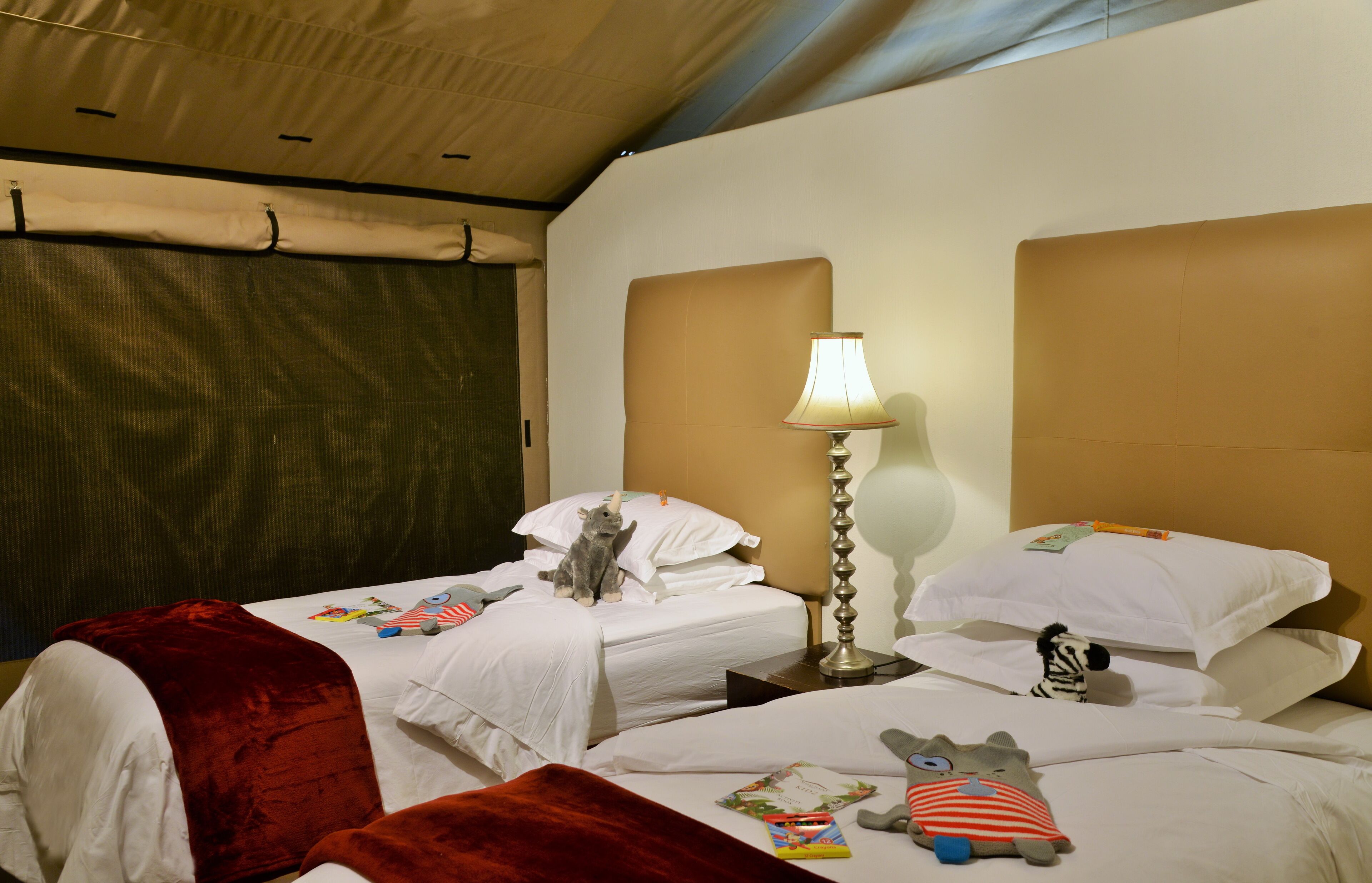 A hotel room with two beds, a red throw blanket, a lamp, and stuffed toys on the beds.
