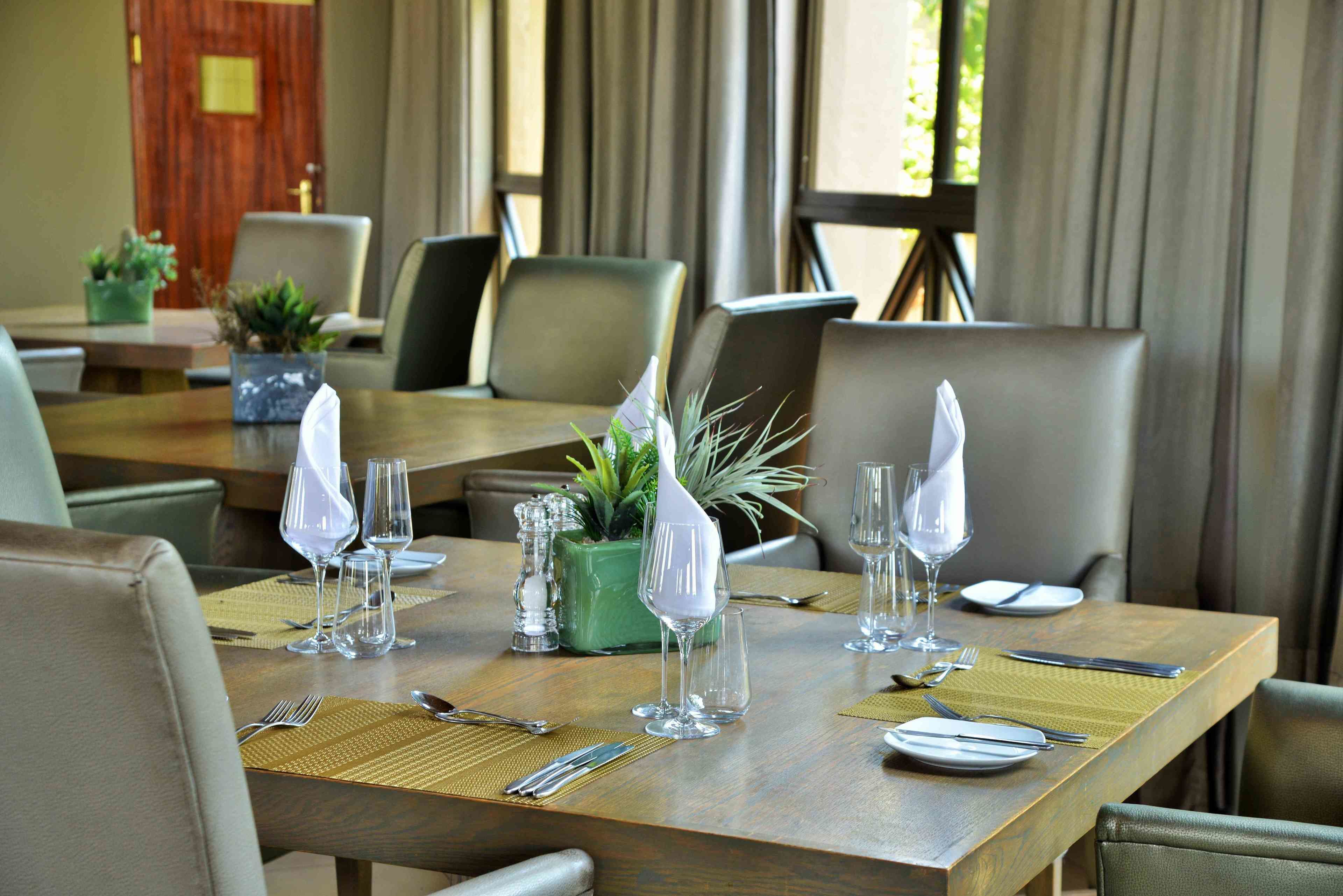 A dining table set with plates, cutlery, and glasses, surrounded by chairs in a well-lit room with a wooden door and curtains in the background.
