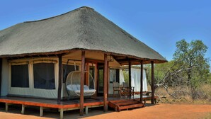 A thatched-roof structure with a covered porch, a hanging chair, and a table with chairs. - Safari Plains (Bela-Bela)