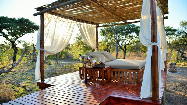 A wooden deck with a canopy, a cushioned seating area, and a table with a fruit basket and a bottle. - Safari Plains (Bela-Bela)