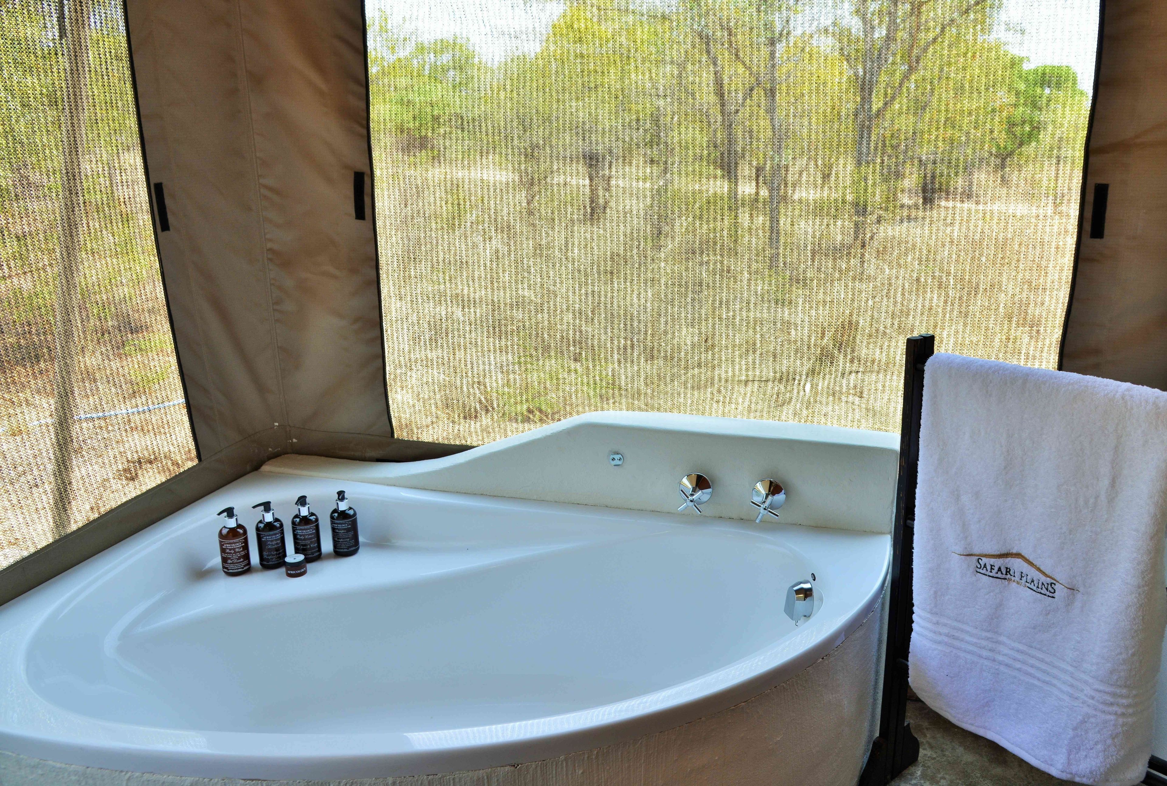 Una bañera grande con una toalla colgada cerca, una ventana con vista a árboles, y una botella de champú.
