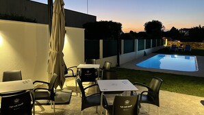 Un espacio exterior con mesas y sillas, una piscina y un atardecer de fondo.