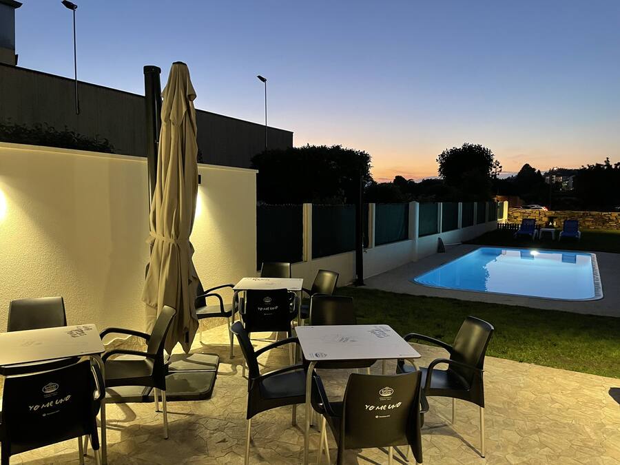 Un espacio exterior con mesas y sillas, una piscina y un atardecer de fondo.