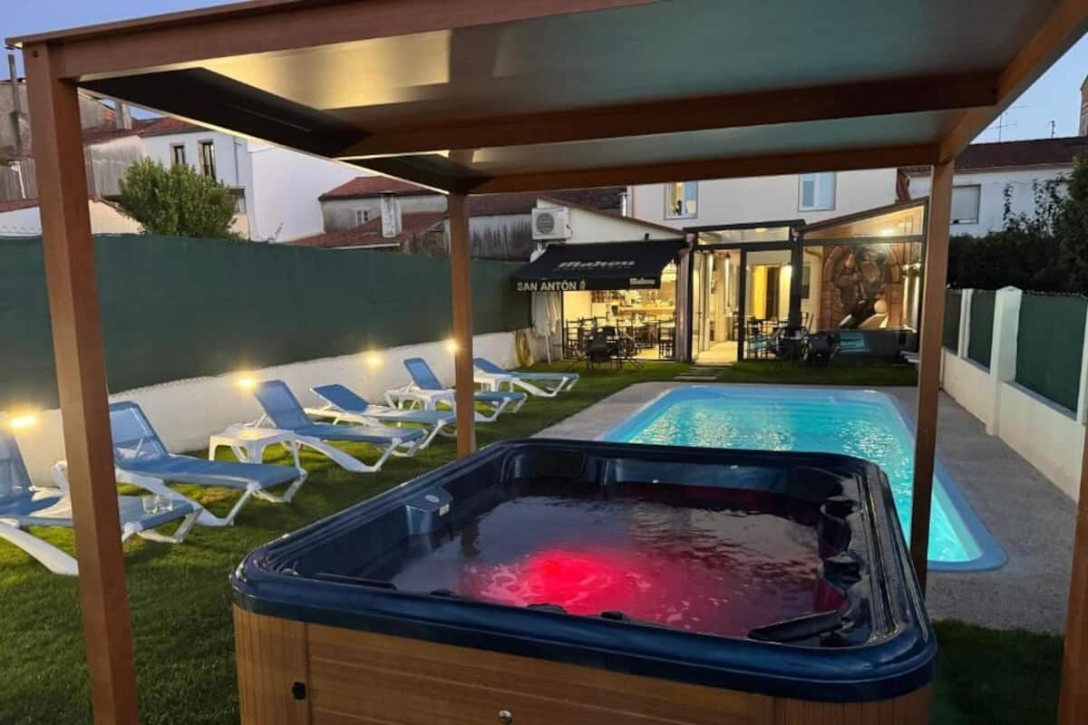 Een hottub onder een houten pergola, een zwembad en ligbedden in een achtertuin.