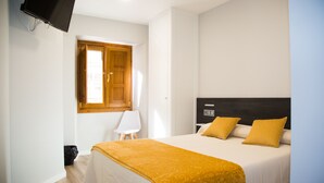Habitación de hotel con cama, televisor montado en la pared, ventana con contraventanas de madera y una silla.