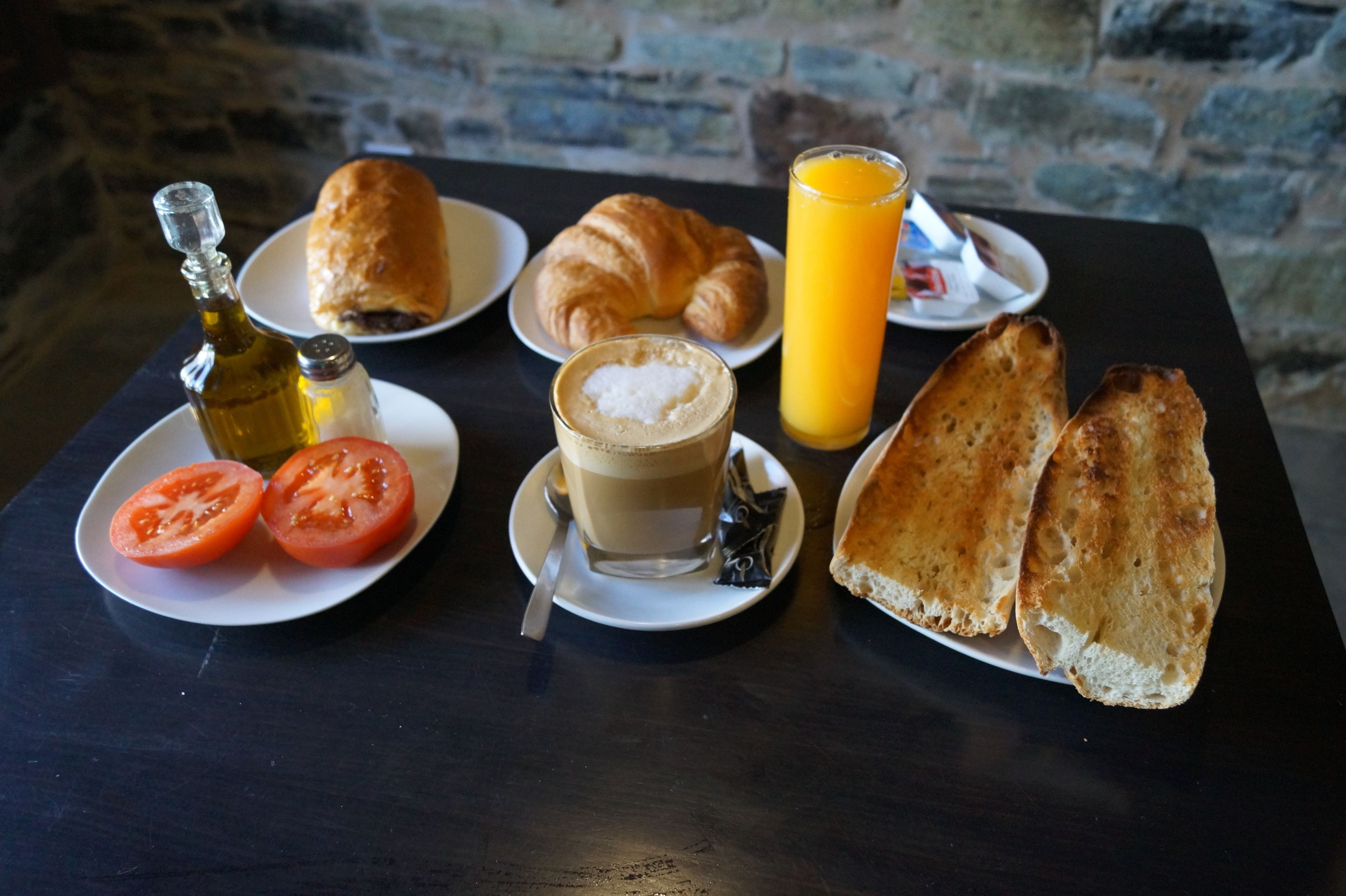A breakfast setting with croissants, toast, tomatoes, a bottle of olive oil, a glass of orange juice, and a cup of coffee on a table.