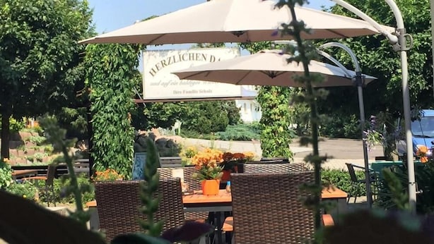 An outdoor seating area with wicker tables and chairs, a large umbrella, and a sign that reads "HERZLICHEN D."