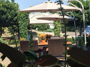 Un espace extérieur aménagé avec des tables et des chaises en osier, un grand parasol et une pancarte sur laquelle on peut lire : « HERZLICHEN D. »