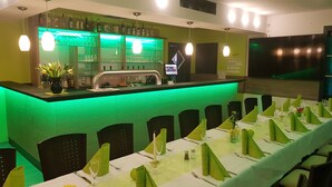 Un restaurante con una zona de bar iluminada en verde, mesas ordenadas con servilletas verdes y un diseño de interiores moderno.