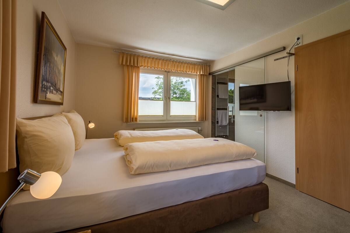 A hotel room with a bed, a TV, a window with curtains, and a painting on the wall.