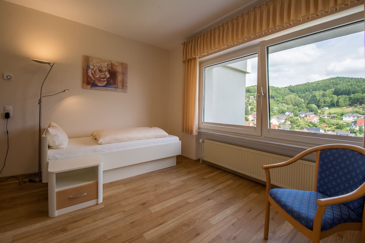 A room with a bed, a chair, a radiator, and a window with a view of a town and hills.