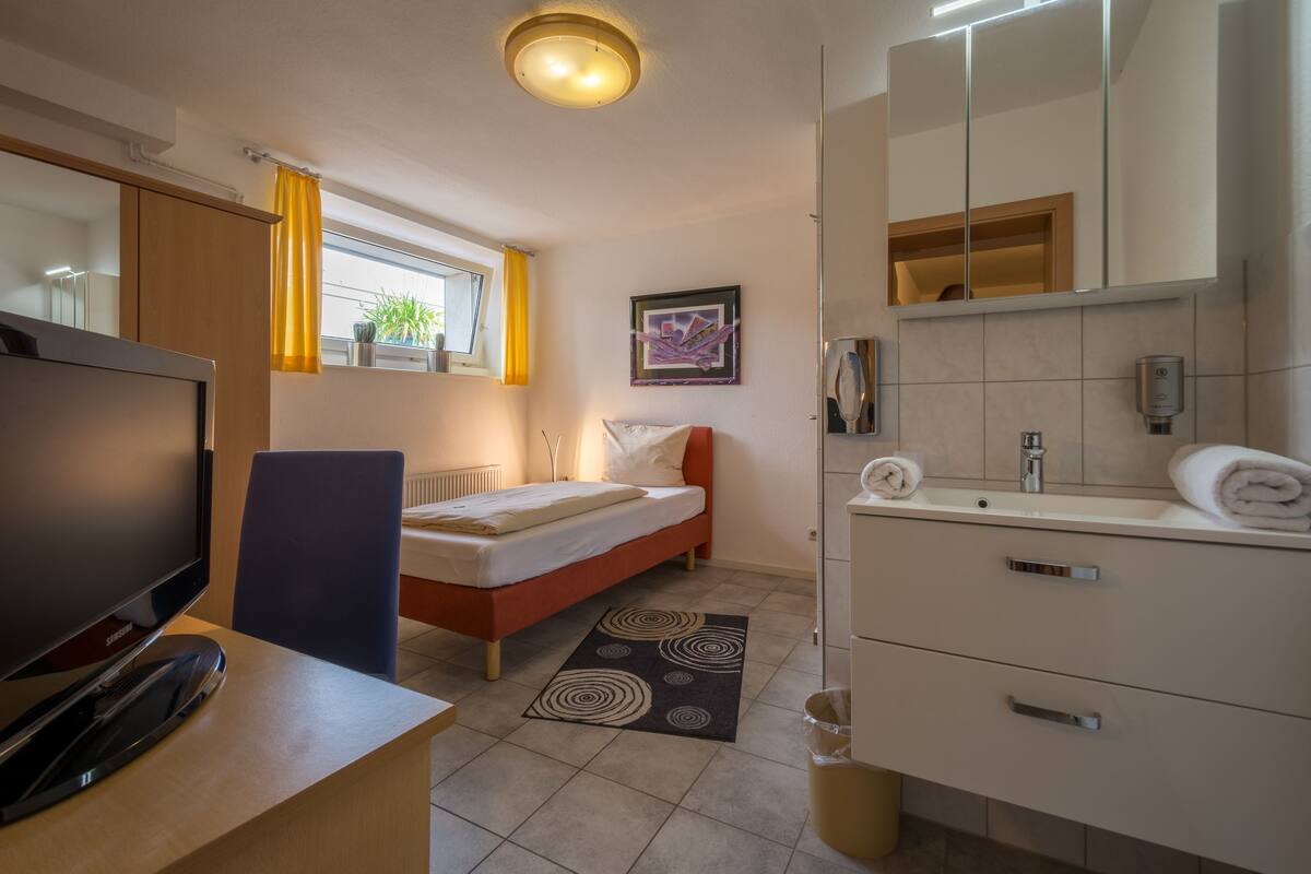 A modern hotel room with a bed, a desk with a computer, a TV, a bathroom with a sink and mirror, and a window with curtains.