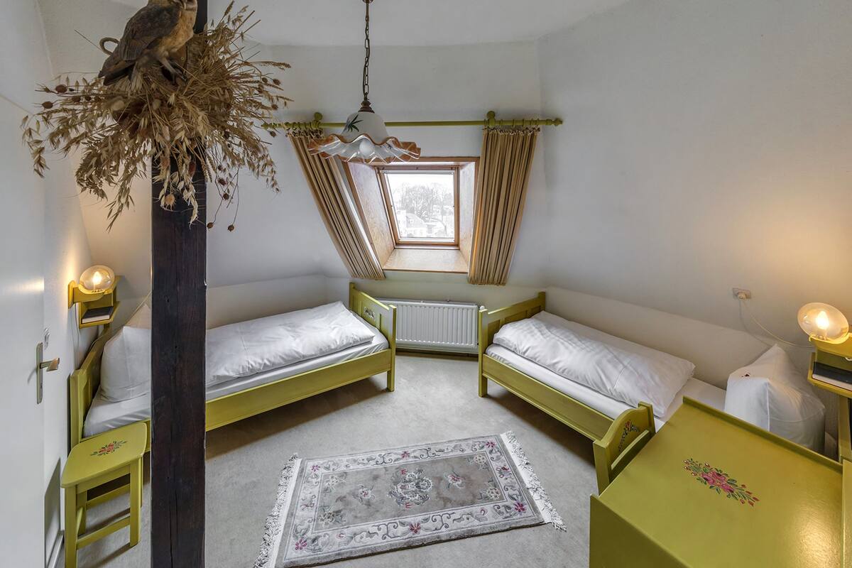 A small, cozy room with two single beds, a window with curtains, a radiator, a small table, and a decorative plant.