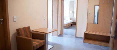 A small, modern room with a single sofa, a small table, and a built-in bench. There is a door leading to a bedroom with a bed and a mirror.