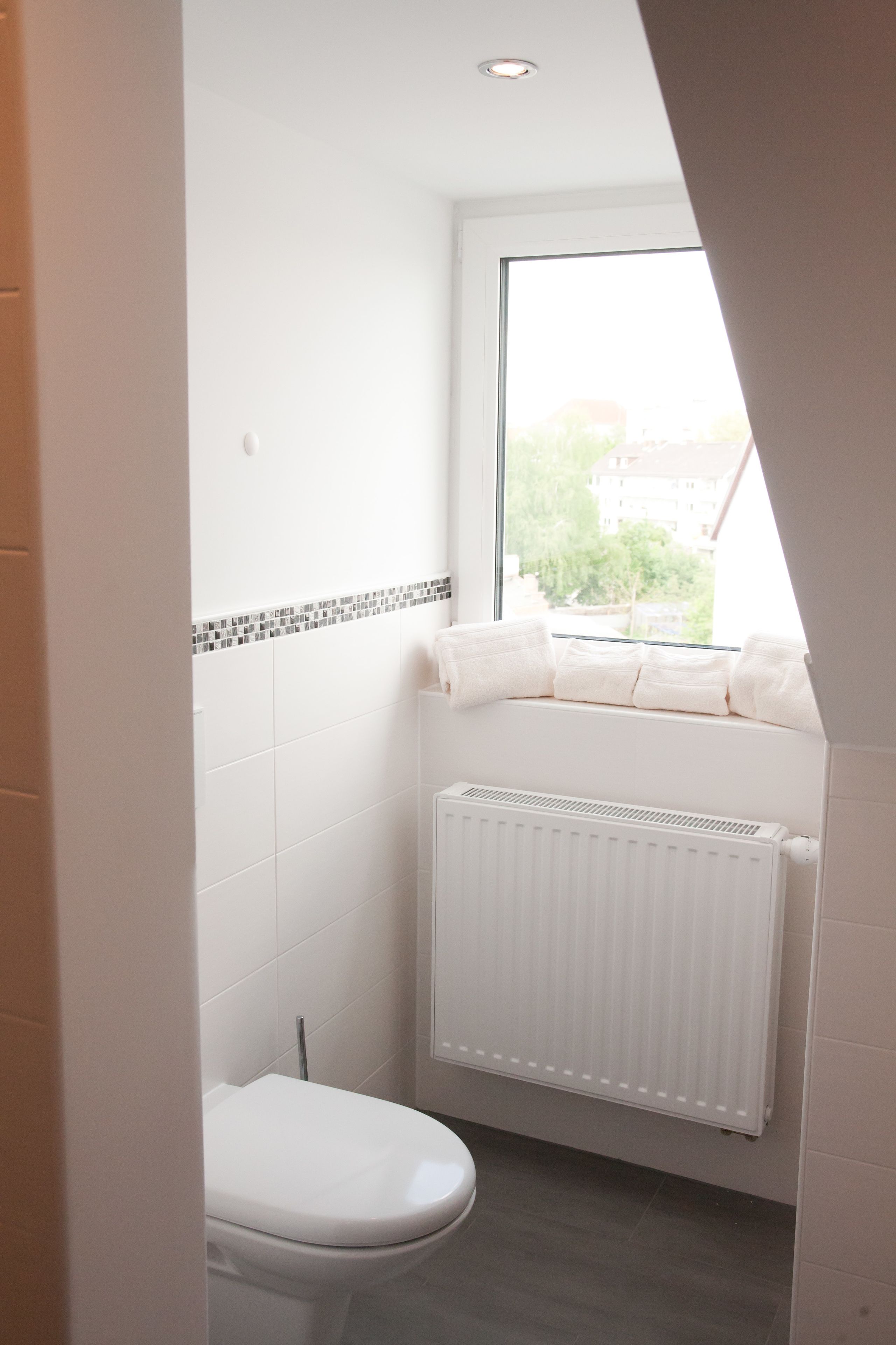 A small, bright bathroom with a toilet, a radiator, and a window with a view of buildings outside.
