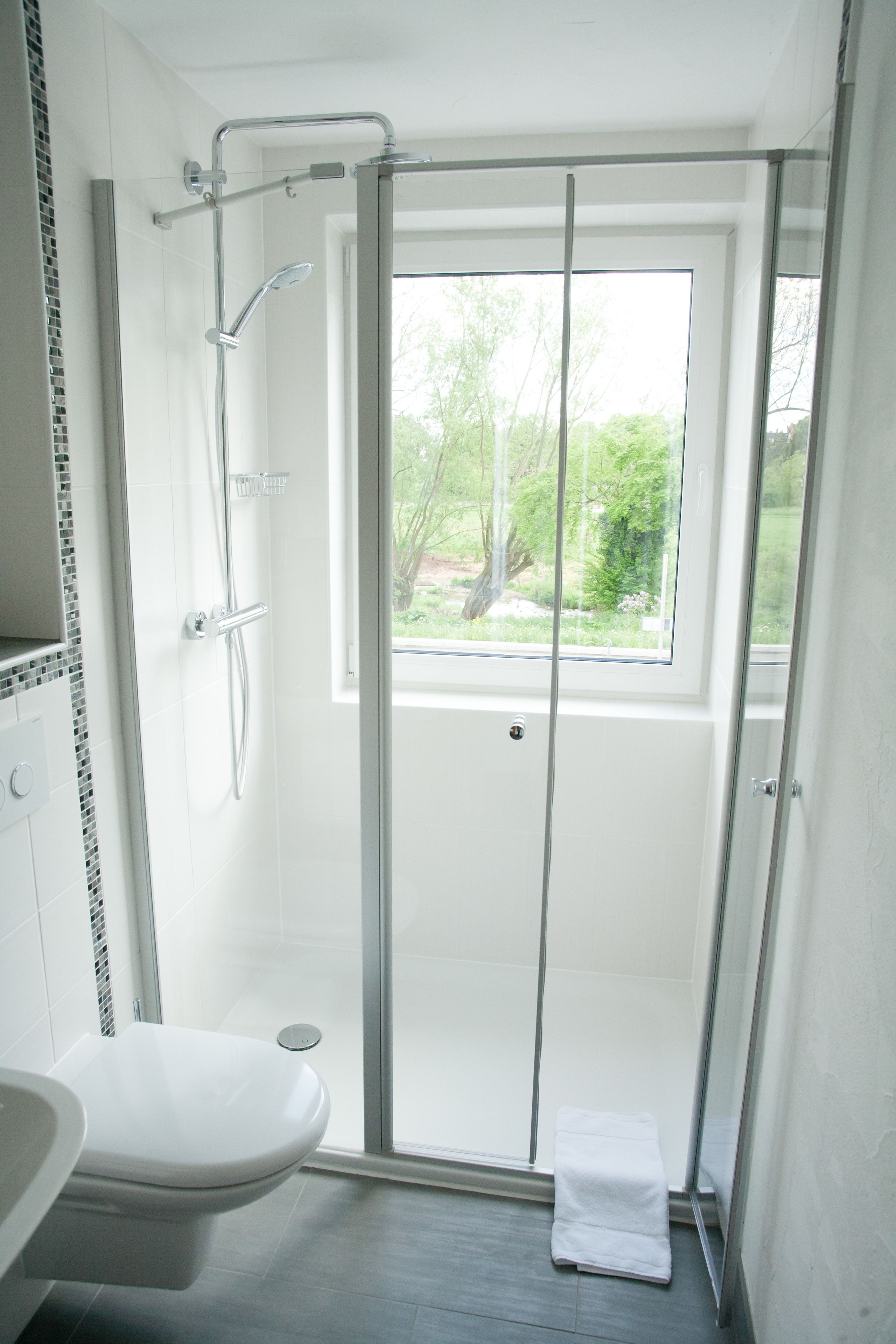 A modern bathroom with a glass-enclosed shower, a toilet, and a window providing a view of greenery outside.