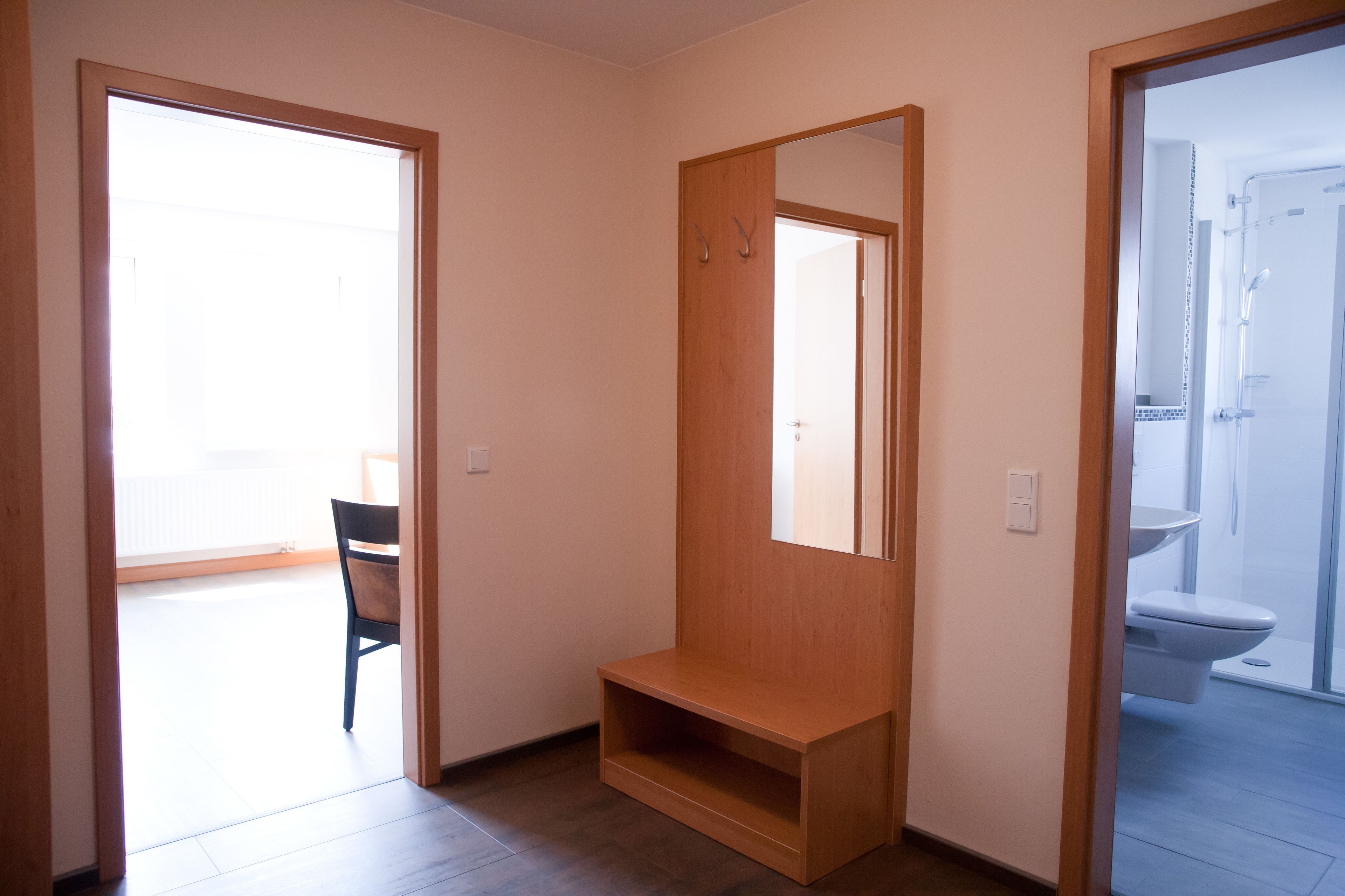 A room with a wooden bench, a mirror, and a door leading to a bathroom with a toilet and shower.