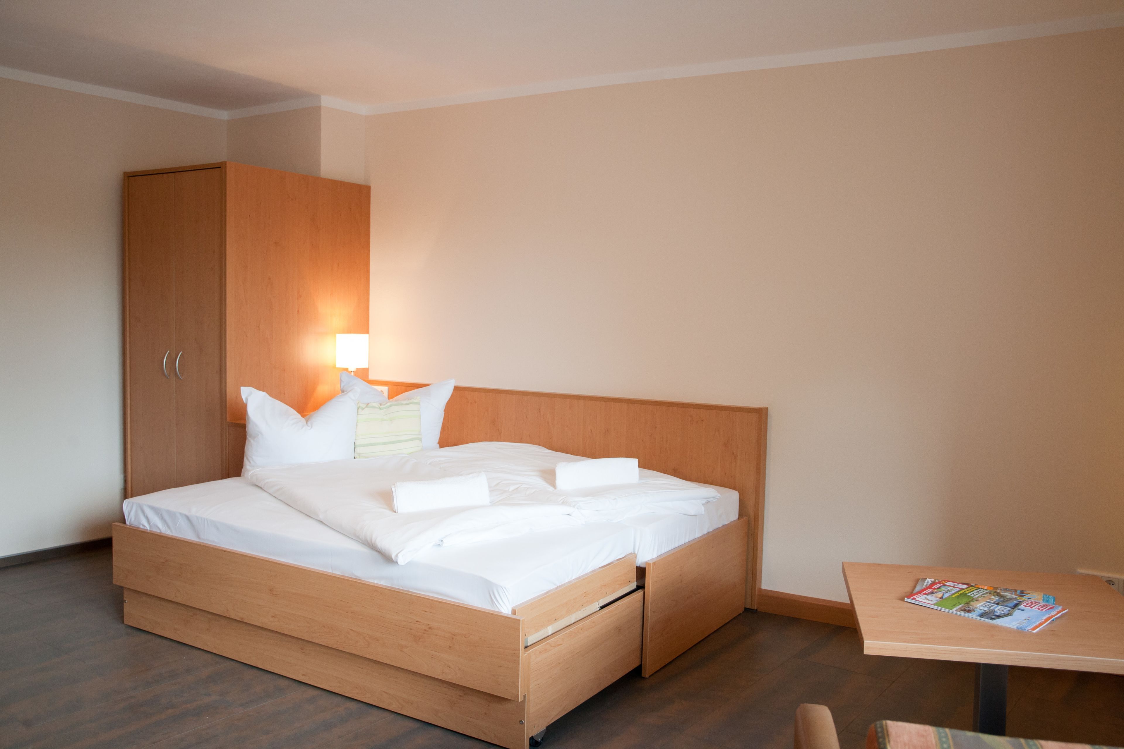 Habitación de hotel con cama de madera, mesita de noche con lámpara, armario de madera y una pequeña mesa con revistas.