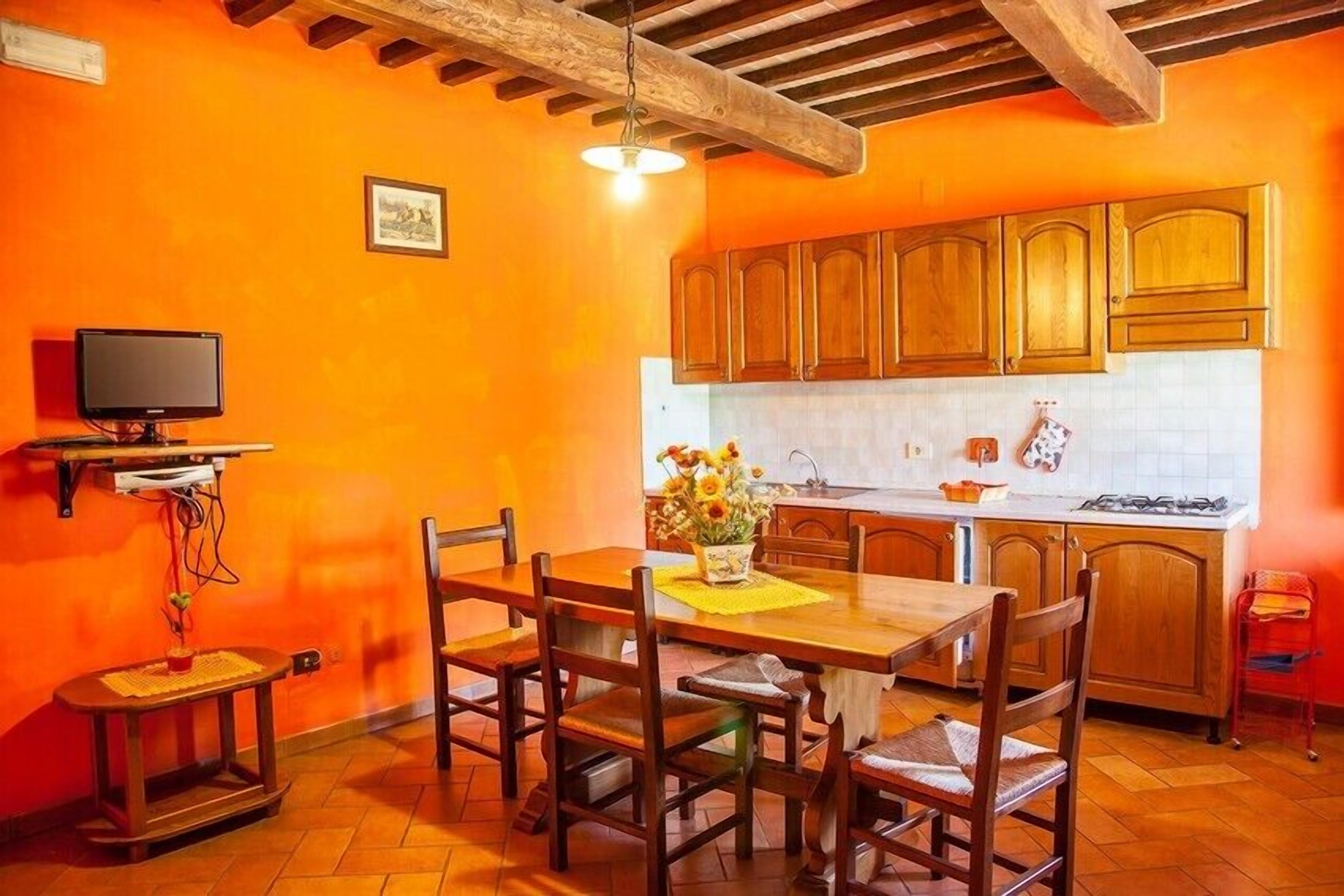 Una cocina con armarios de madera, una mesa de comedor con sillas, un televisor en un estante y un jarrón sobre la mesa.