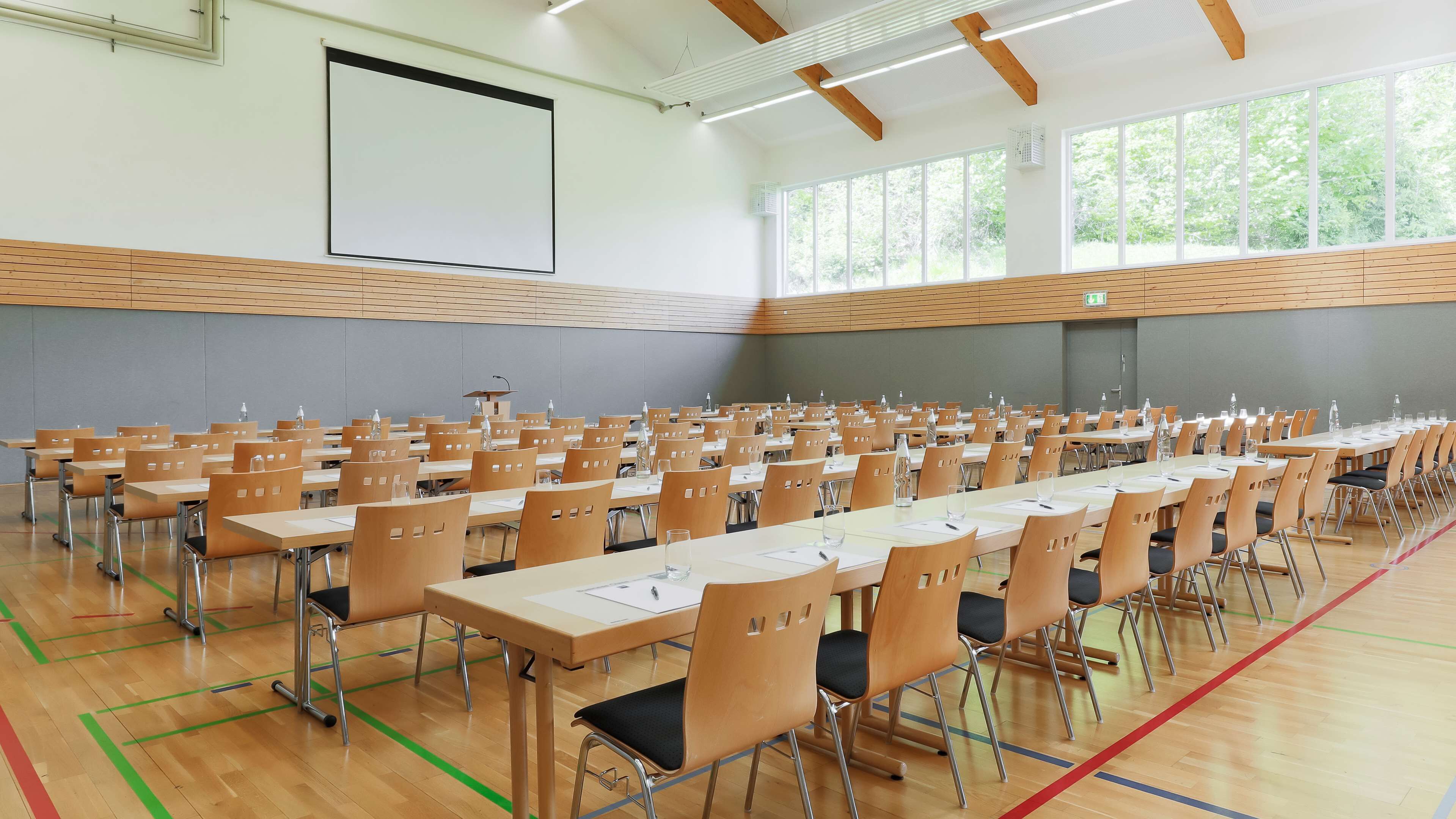 Une grande salle de conférence avec des rangées de tables et de chaises, un écran de projection et de grandes fenêtres.