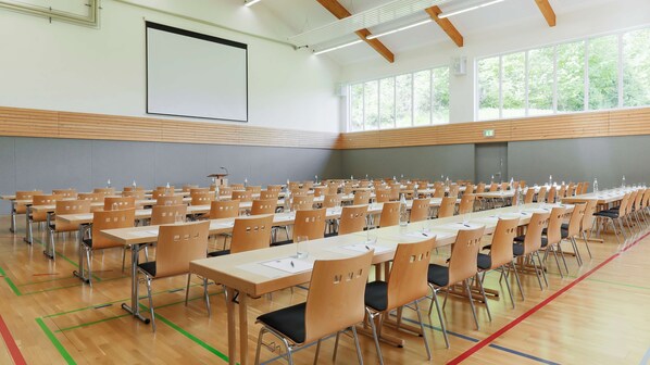 Une grande salle de conférence avec des rangées de tables et de chaises, un écran de projection et de grandes fenêtres.