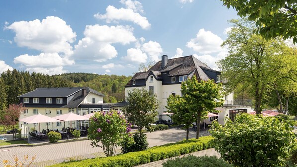 Ein Hotel mit einem gepflasterten Innenhof, Sitzgelegenheiten im Freien und Blick auf einen bewaldeten Hang.