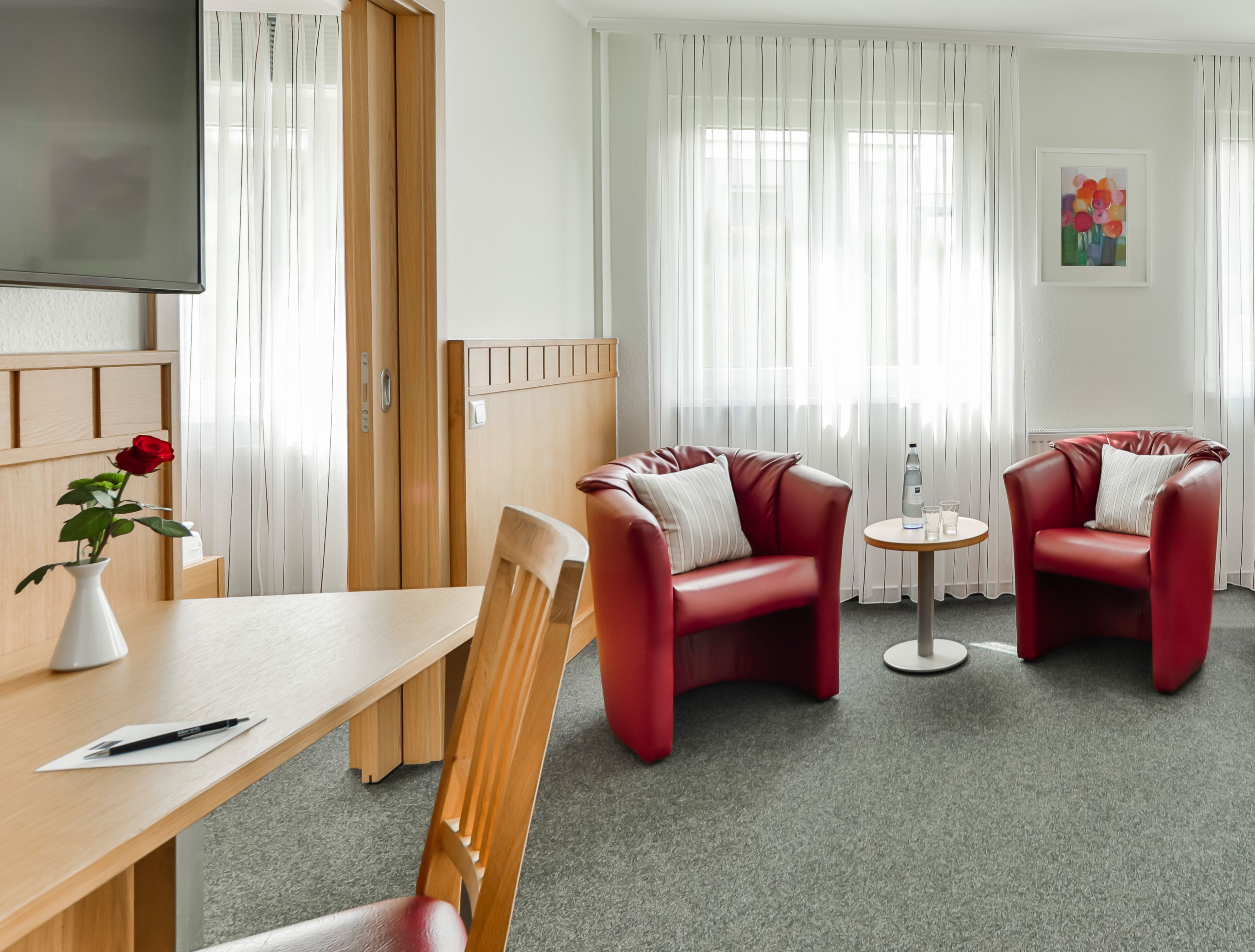 A modern hotel room with a wooden desk, two red leather sofas, a small round table, and a TV on the wall.
